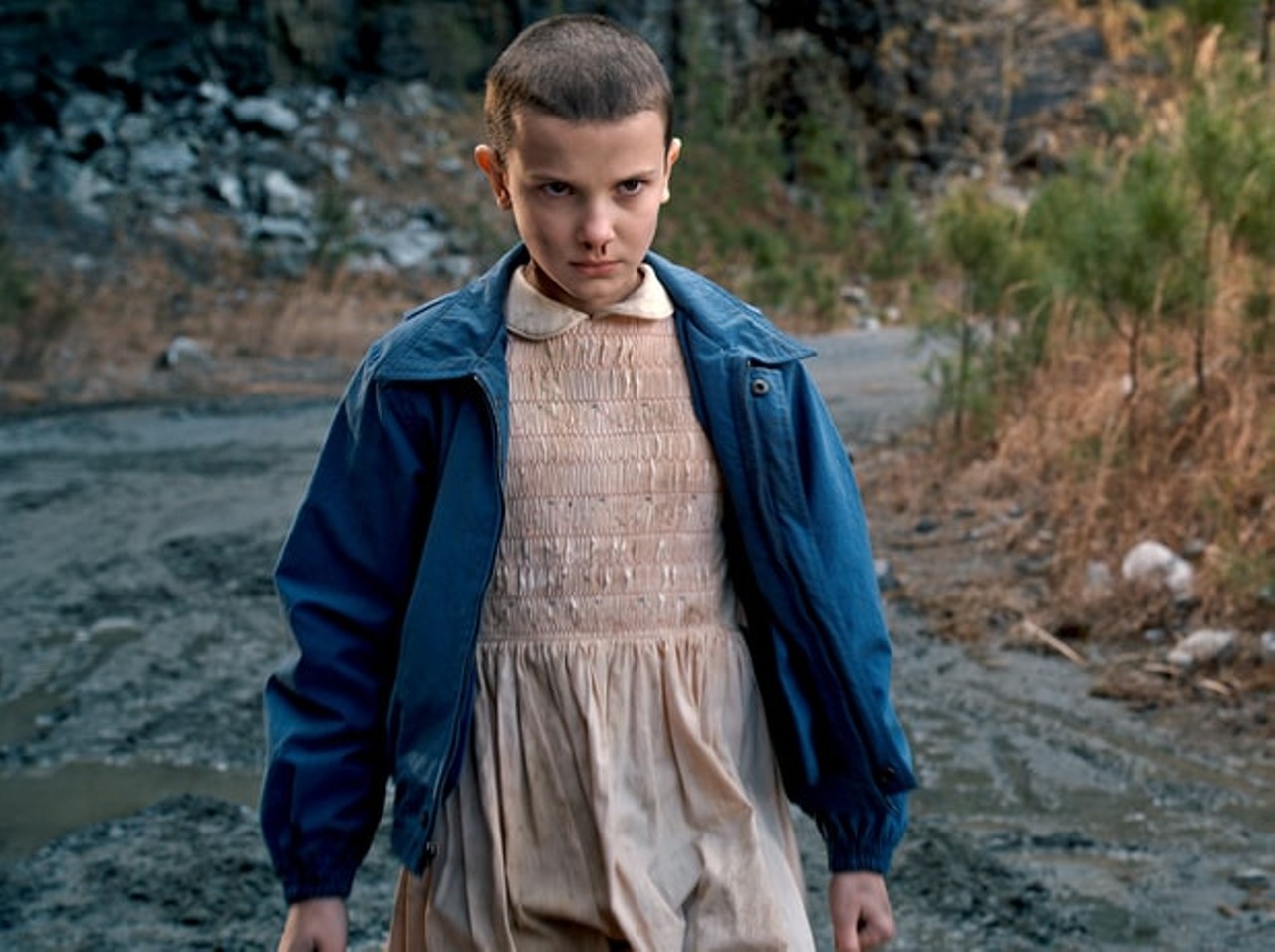 To be Eleven (aka the best Stranger Things character) you need a Short Blonde wig, a pink ruffed dress with a white collar, a navy bomber jacket, a box of Eggo's, knee high white striped socks, and white sneakers. 
Photo courtesy of Netflix 