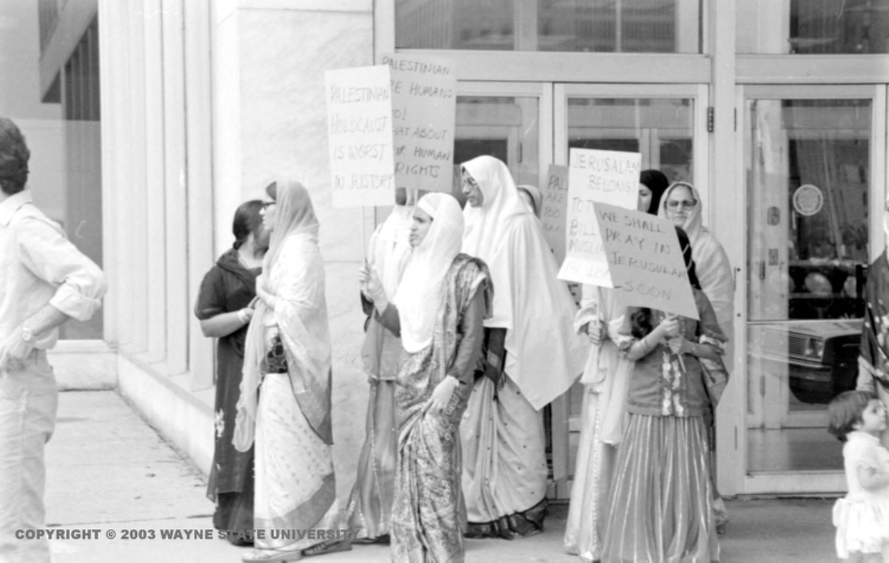 1970s | Women wearing traditional clothing of long skirts, shawls and head scarves stand together holding signs, one reads, "We shall pray in Jerusalem, soon," on sidewalk in front of unidentified building.