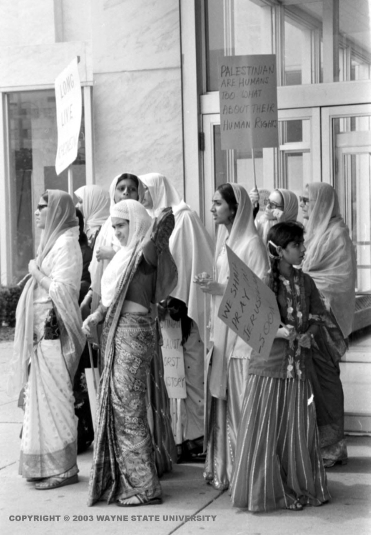 1970s | Women wearing saris and head scarves stand together holding signs, one reads, "Palestinians are human too, what about their human rights," on steps of unidentified building.