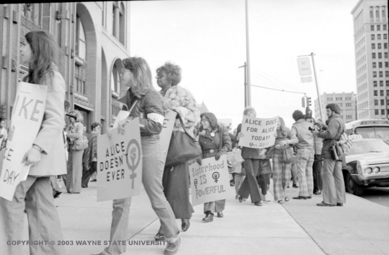 1970s | Group of women with signs march on sidewalk near building, participating in Alice Doesn't Day, a one-day work stoppage organized by the National Organization for Women to draw attention to the work that women perform in society.