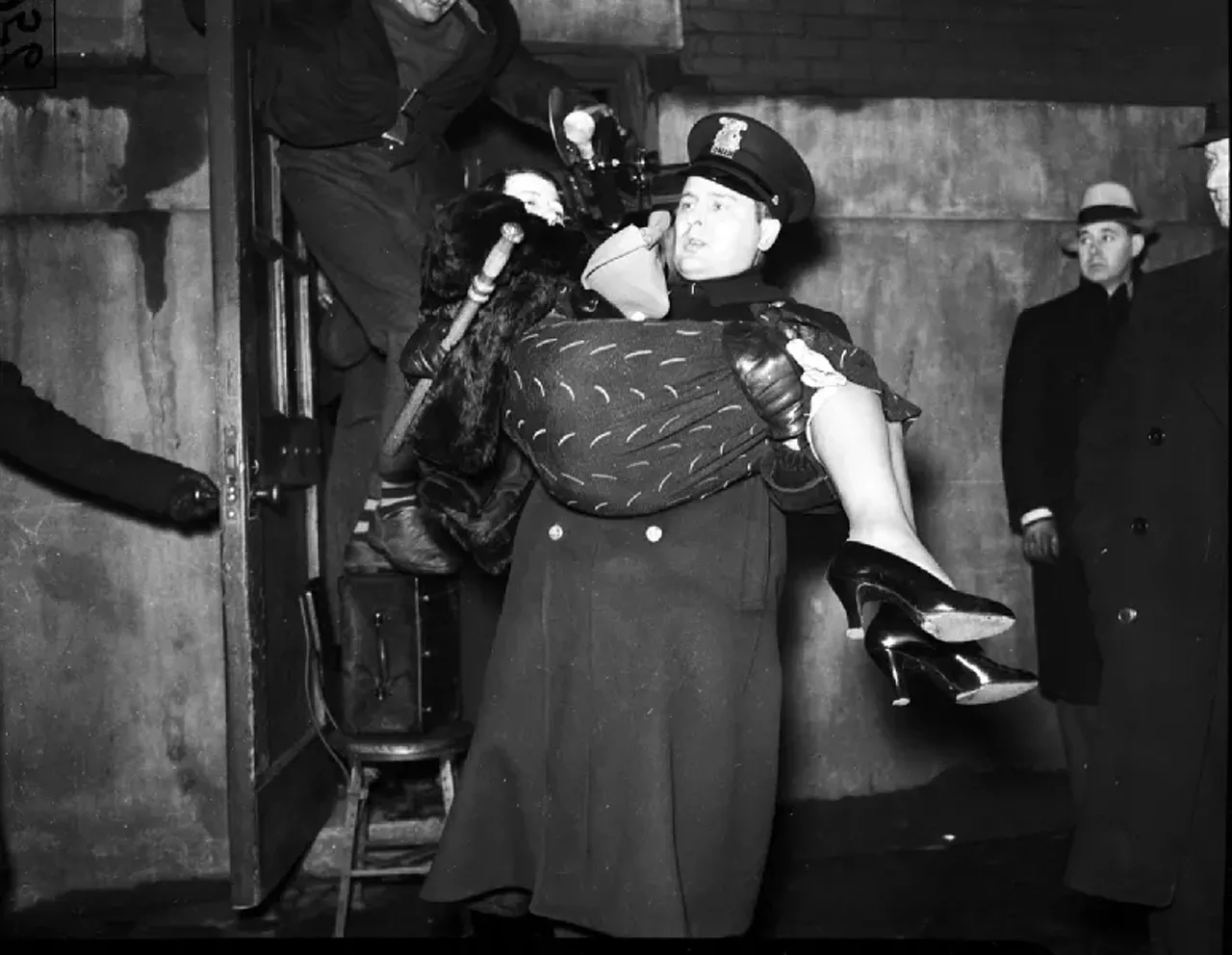 1930s | Uniformed police officer carries woman (striking employee?) in his arms, during sit-down strike at Mazer-Cressman Cigar Company in Detroit, Michigan.