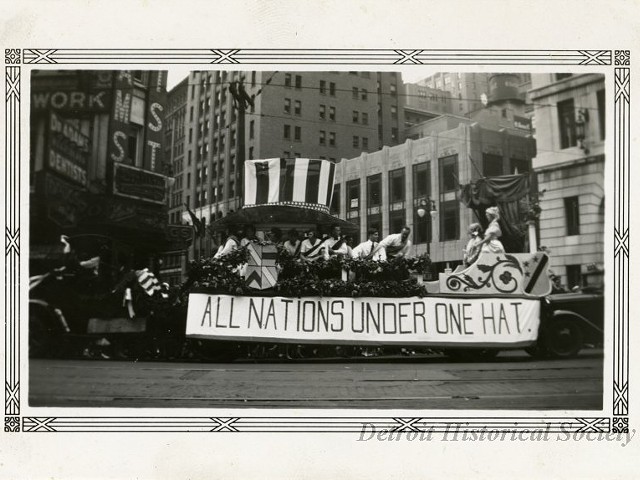 "Black and white photographic print with a decorative border taken at the 1932 Fourth of July parade on Michigan Avenue at Griswold Street facing north. A parade float consisting of a large Uncle Sam hat above a group of people wearing sashes, and an ornate carriage at the end where a man and woman in fancy costume stand is being pulled west along Michigan Avenue behind a truck. A banner reading, "All Nations Under One Hat," is hung from the side of the float. The Peoples Wayne County Bank, David Stott Building, and Dr. Adams' dentist office are in the background. "Jul 4 1932," and "108," are stamped on the verso."