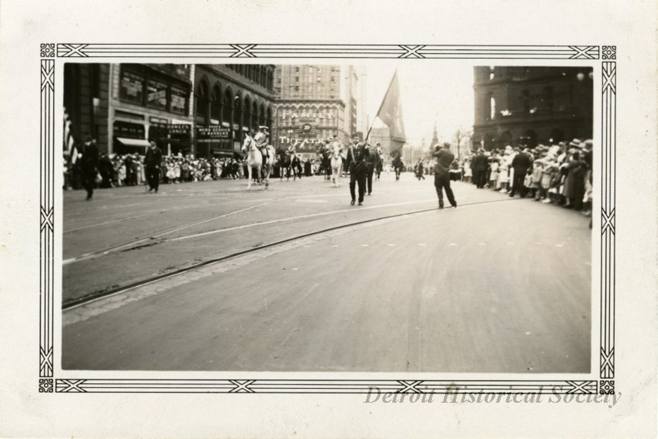 "Black and white photographic print with a decorative border taken at the 1932 Fourth of July parade on Michigan Avenue between Shelby Street and Griswold Street facing east. A group of men wearing Revolutionary War costume on horseback, surrounded by men in contemporary military dress proceed west along Michigan. A photographer is taking of picture of one of the men in modern uniform, who holds a flag on which "George Washington" is embroidered. A crowd lines both sides of Michigan. Bowles Lunch, and Rose's Men's Service barbershop are among the storefronts on the north side of Michigan. The Family Theatre, City Hall, Cadillac Square Building, the Michigan Soldiers' and Sailors' Monument, and the Wayne County Building are all visible in the background. "Jul 4 1932," and "108," are stamped on the verso."
