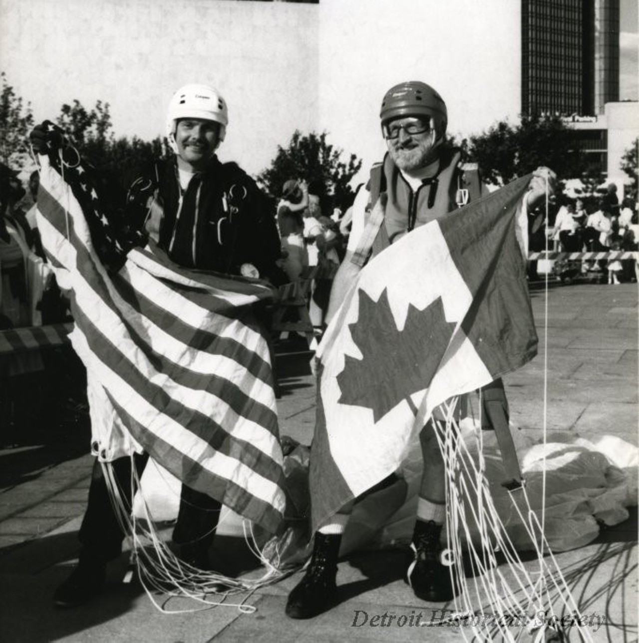 "Black and white photographic print depicting two men standing in Hart Plaza holding an American and Canadian flag, wearing helmets and parachutes. In the background are the Renaissance Center, Ford Auditorium and many people.
Caption reads: Skydivers carrying American and Canadian flags are symbolic of the 24 year-old Detroit/Windsor International Freedom Festival. Their free fall and landing on Hart Plaza is part of the International Air and Water Show on June 25 and June 26. The Freedom Festival is a ten day celebration of friendship and freedom between the United States and Canada. It features more than 50 events and runs from Friday, June 25 through Sunday, July 4.
Contact: Sharlan Douglas"