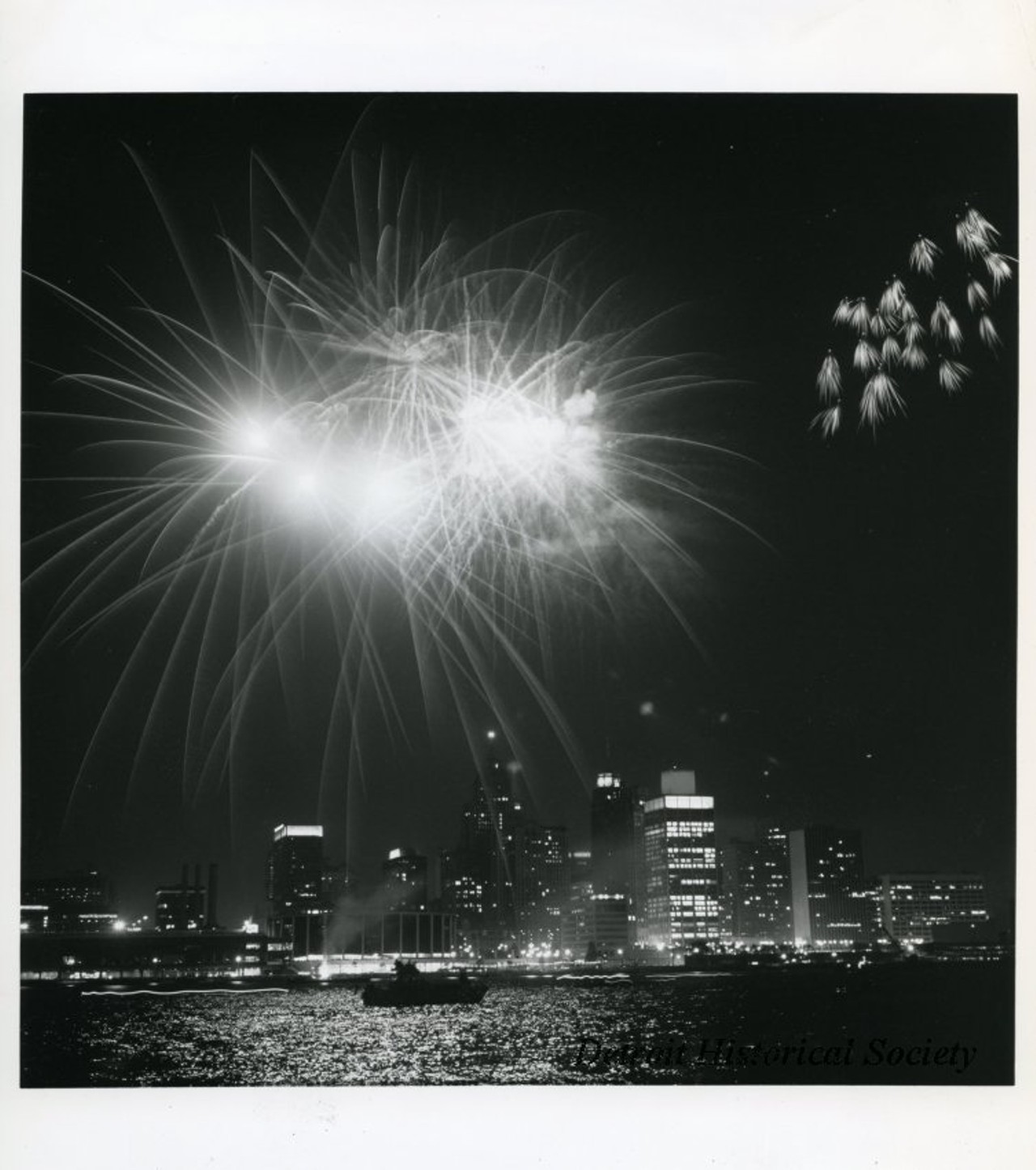 "Black and white photographic print depicting a fireworks display over the Detroit River at night, with the downtown skyline in the background. In view are Cobo Center, Detroit Bank and Trust Tower, One Woodward Avenue, and City-County Building."