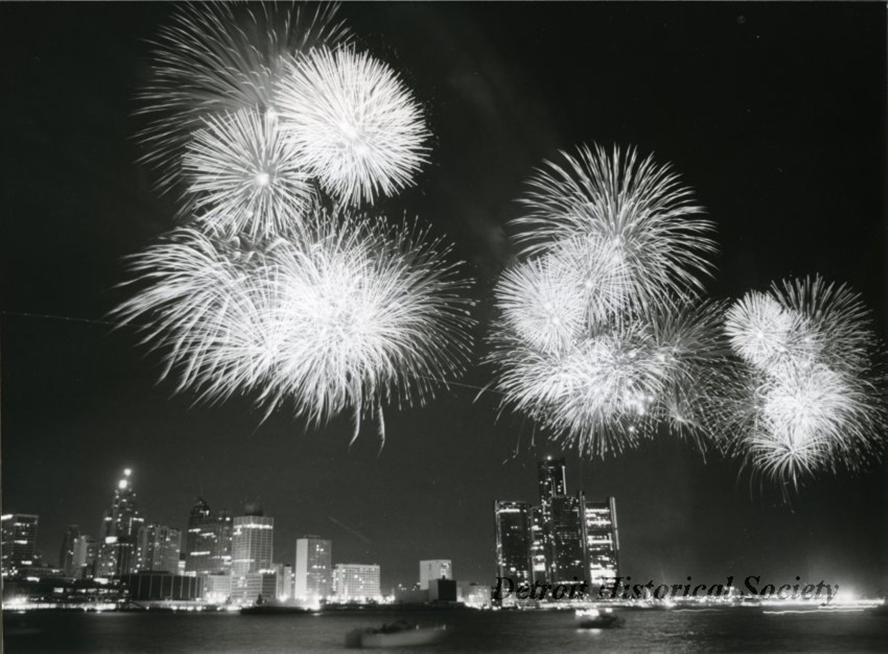 "Black and white photographic print depicting a fireworks display over the Detroit River at night, with the downtown skyline in the background. In view are the Renaissance Center, Cobo Arena, Guardian Building, and City-County Building.
Caption reads: International skies will light up as more than eight tons of fireworks are launched from the Detroit River on June 30 at the 24th International Freedom Festival. The largest fireworks display in North America is sponsored by the J. L. Hudson Co., the Stroh Brewery Co., and WDIV TV-Channel 4.
Contact: Sharlan Douglas"