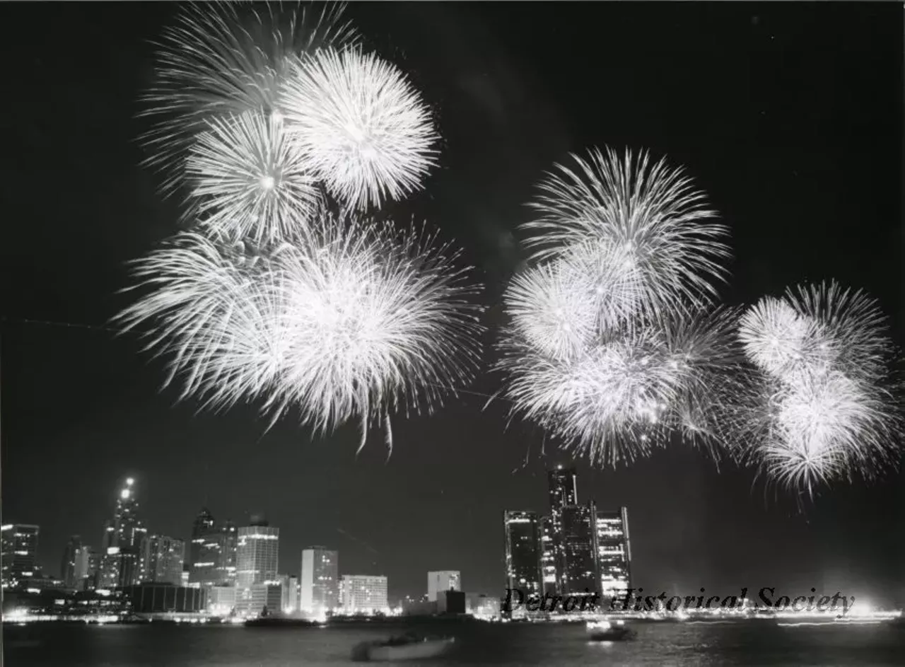 "Black and white photographic print depicting a fireworks display over the Detroit River at night, with the downtown skyline in the background. In view are the Renaissance Center, Cobo Arena, Guardian Building, and City-County Building. Caption reads: International skies will light up as more than eight tons of fireworks are launched from the Detroit River on June 30 at the 24th International Freedom Festival. The largest fireworks display in North America is sponsored by the J. L. Hudson Co., the Stroh Brewery Co., and WDIV TV-Channel 4. Contact: Sharlan Douglas"