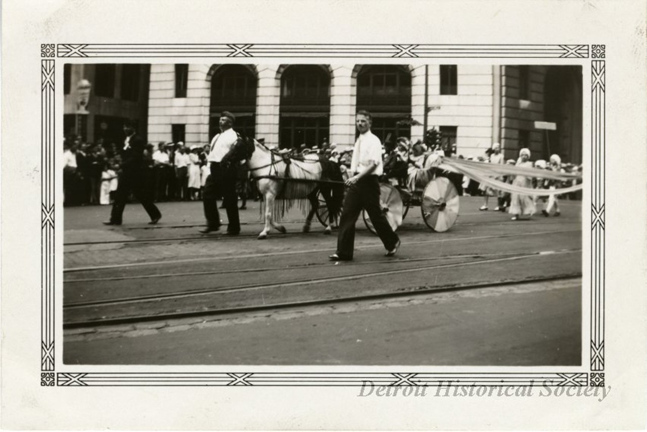 "Black and white photographic print with a decorative border taken at the 1932 Fourth of July parade on Michigan Avenue at Griswold Street facing northeast. Three men walk alongside a horse-drawn carriage carrying a man and two children, heading west along Michigan. A group of children follow behind the carriage. A crowd stands in the background along Michigan in front of the Peoples Wayne County Bank. "Jul 4 1932," and "108," are stamped on the verso."