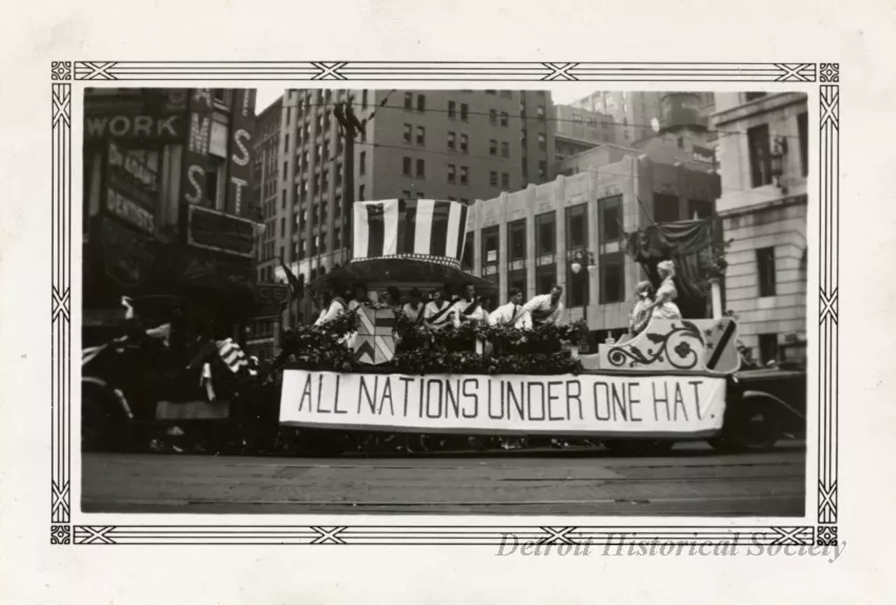 "Black and white photographic print with a decorative border taken at the 1932 Fourth of July parade on Michigan Avenue at Griswold Street facing north. A parade float consisting of a large Uncle Sam hat above a group of people wearing sashes, and an ornate carriage at the end where a man and woman in fancy costume stand is being pulled west along Michigan Avenue behind a truck. A banner reading, "All Nations Under One Hat," is hung from the side of the float. The Peoples Wayne County Bank, David Stott Building, and Dr. Adams' dentist office are in the background. "Jul 4 1932," and "108," are stamped on the verso."