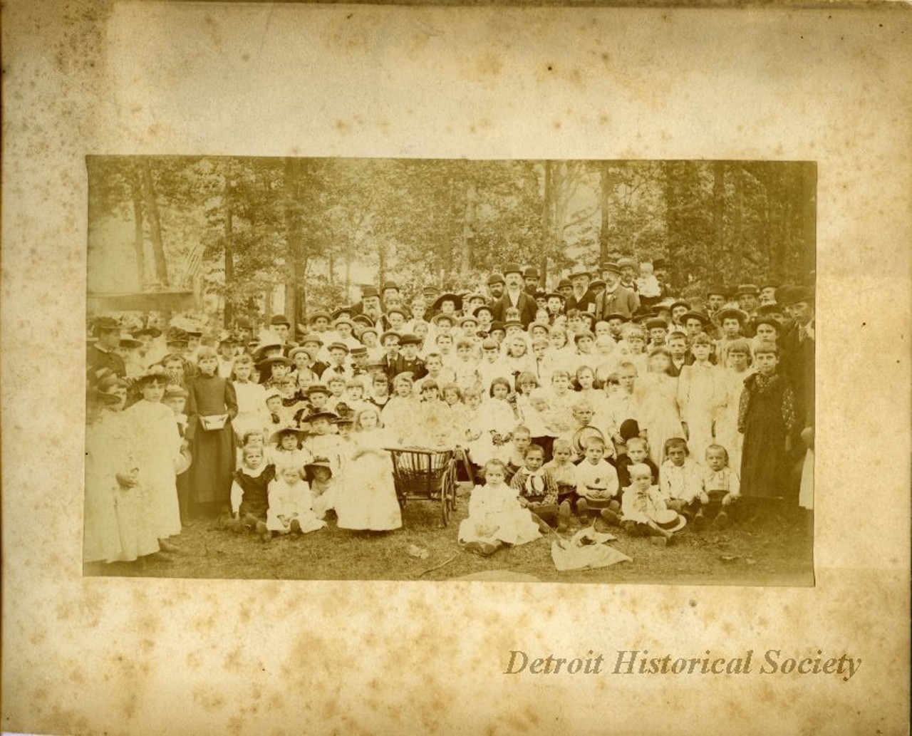 "One sepia-toned photograph of the congregation of St. James Lutheran Church in Detroit. The photo is mounted on white matte board and shows a large group of men, women, and children who are standing or seated in a park. All are well-dressed in Sunday attire. A grassy lawn is visible in the foreground and tall trees can be seen in the background. A middle-aged man with a moustache and bowler hat in the right center background may be church pastor, Rev. August G. Bergener, although he does not appear to be wearing a clerical collar. A handwritten note attached to the photo reads 'Probably a picture taken at the annual picnic at Clark Park. The group marched over to the park every year on the Fourth of July for this event in the early years.'"