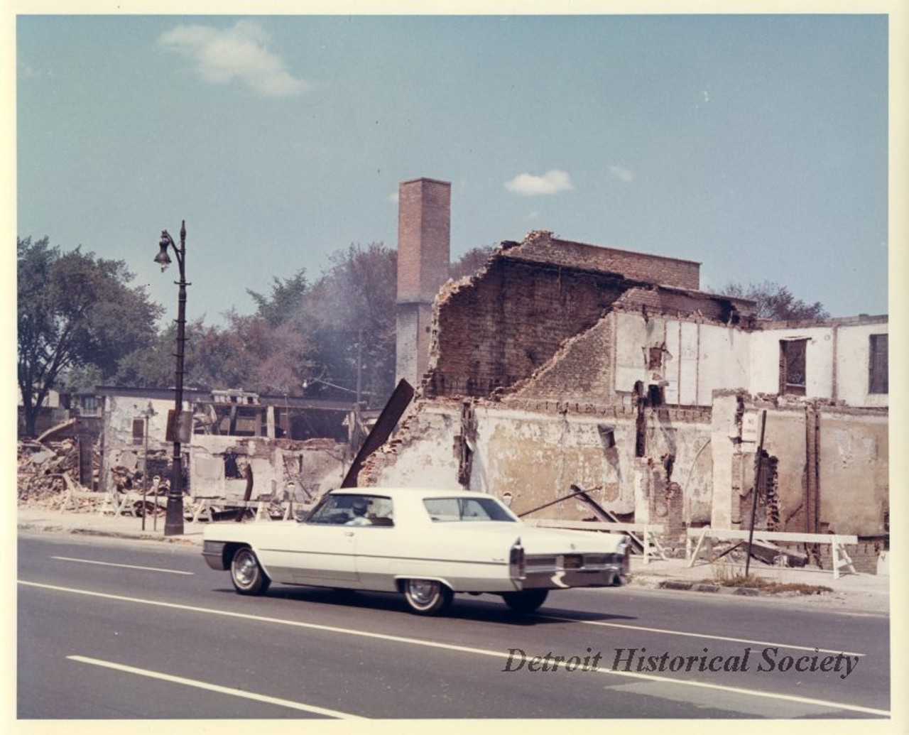 "One color photograph of a row of buildings that were burned down during the 1967 Detroit Riot. A white Cadillac is driving along the street in the foreground. A red ink stamp on the verso shows 'D.F.D. [Detroit Fire Department] Photo, by Capt. J. A. Mancinelli.'"