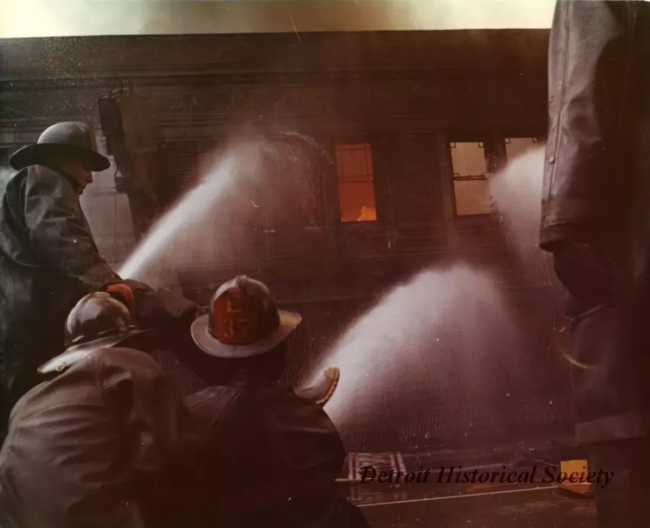 "One color photograph of a group of four Detroit Fire Department firemen who are trying to extinguish a blaze at a row of buildings located on 12th Street between Taylor and Clairmount Streets. The buildings had been set on fire during the 1967 Detroit Riot. A red ink stamp on the verso shows 'D.F.D. [Detroit Fire Department] Photo, by Insp. Barney Wasowicz.'"