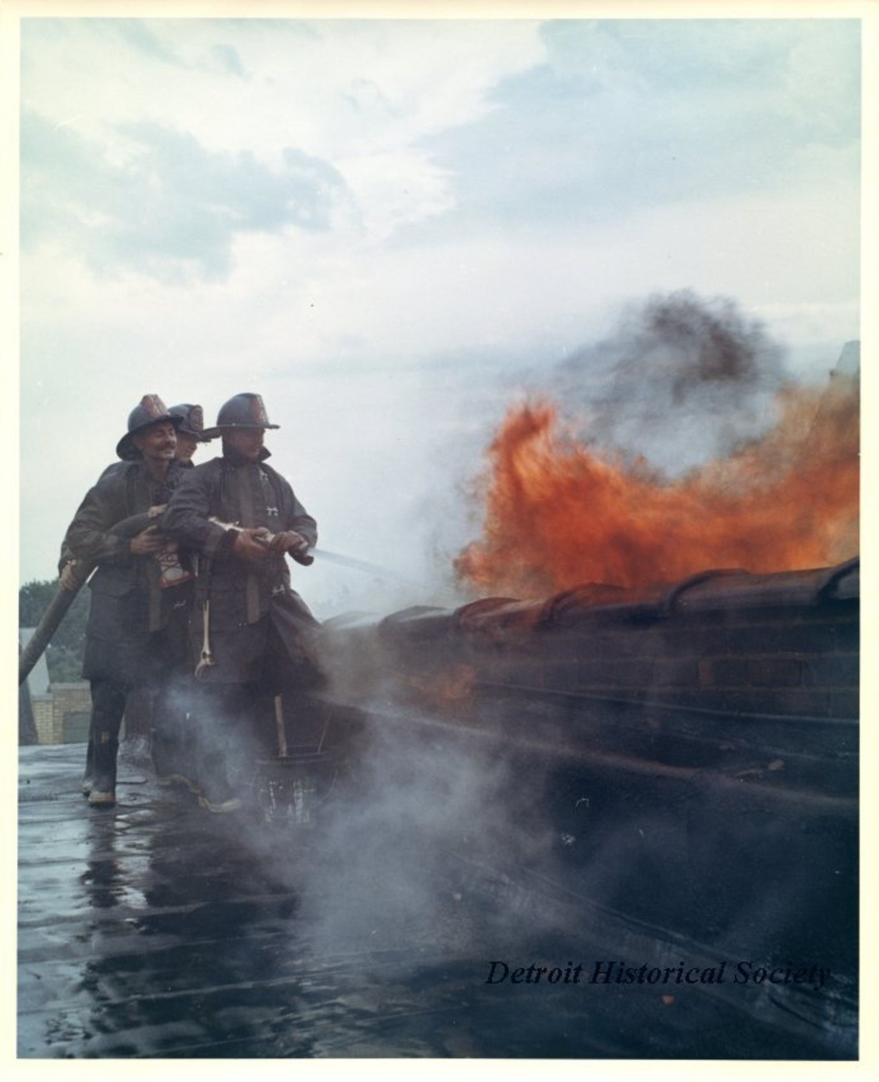 "One color photograph of a group of three Detroit Fire Department firemen who are trying to extinguish a blaze from the roof of a row of buildings located on 12th Street between Taylor and Clairmount Streets. The buildings had been set on fire during the 1967 Detroit Riot. A red ink stamp on the verso shows 'D.F.D. [Detroit Fire Department] Photo, by Insp. Barney Wasowicz.'"