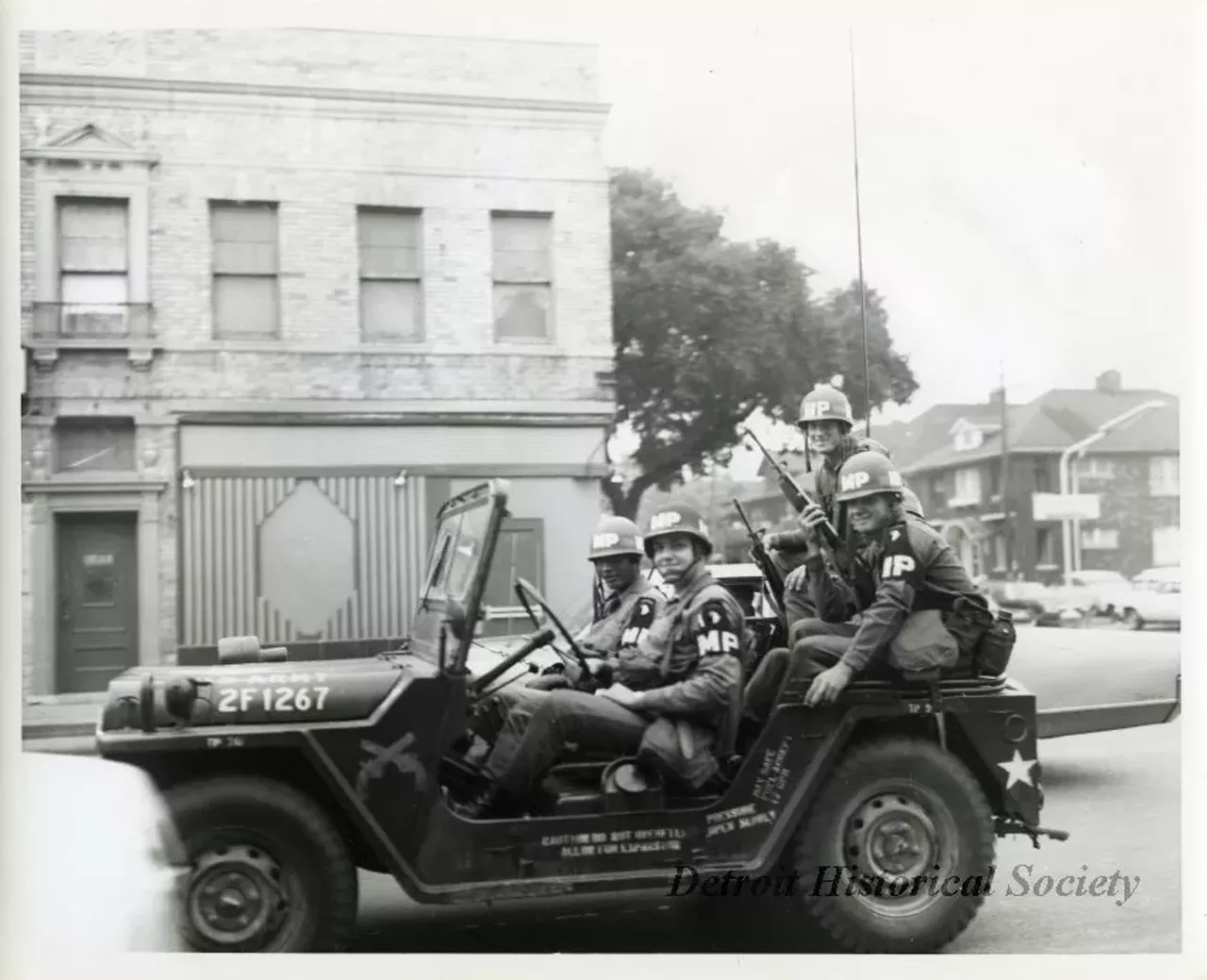 "One black and white photograph of four MP (military police) soldiers who are on patrol in a jeep during the 1967 Detroit Riot. Two soldiers, who are seated in the back of the jeep, are carrying rifles. A 2-story commercial building is visible in the left background. Several houses and parked cars can be seen in the right background."