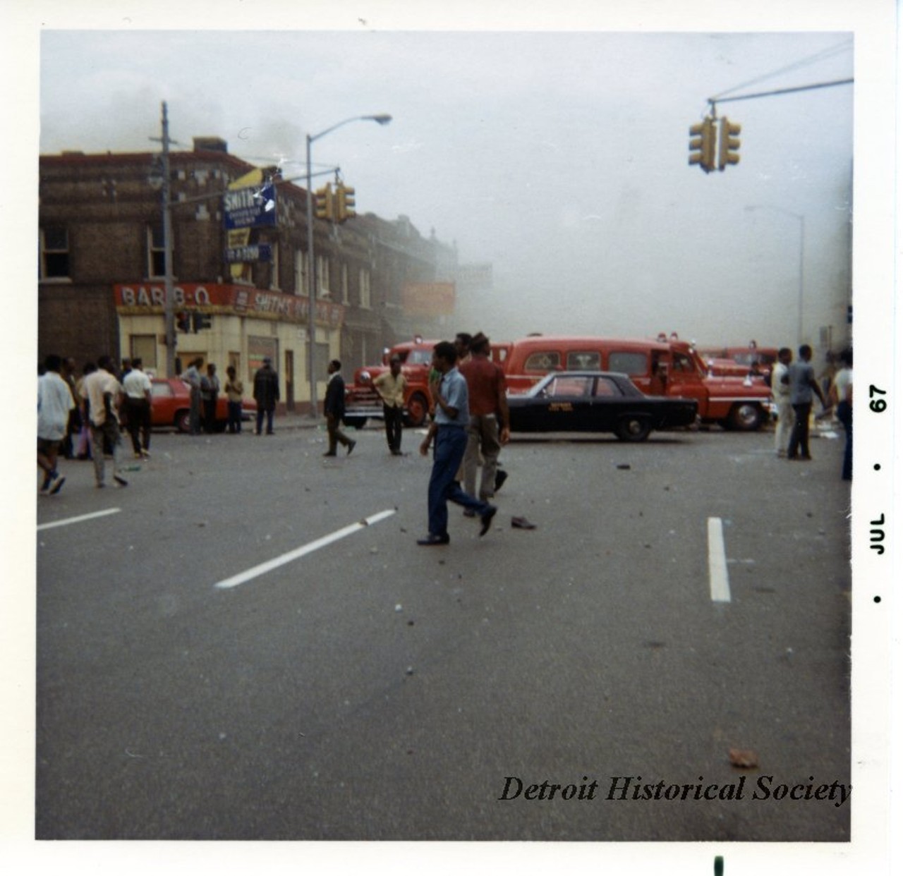 "One color photograph of the intersection of 12th and Clairmount Streets during the 1967 Detroit Riot. The photo shows many police and fire vehicles parked in the street intersection with a large number of bystanders who are viewing the scene. Smith's Bar-B-Q Restaurant, located at 9050 12th Street, can be seen in the left background. Smoke is visible in the center and right background areas of the photo. A printed black date stamp on the edge of the photo shows 'Jul., 67.'"