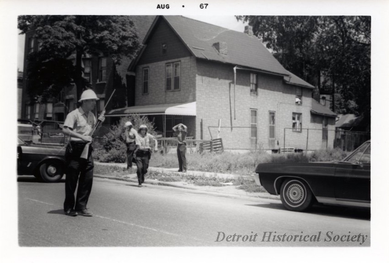 "One digital scan of a black and white photograph which shows a young white man being stopped by police during the 1967 Detroit Riot.  The man is standing on a sidewalk on a residential street and has his hands on his head. Two police officers are walking away from him and another office is standing in the street and is holding a rifle. A police car is parked on the street in the left foreground and a car is parked in the right foreground. A house and vacant lot can be seen in the background."