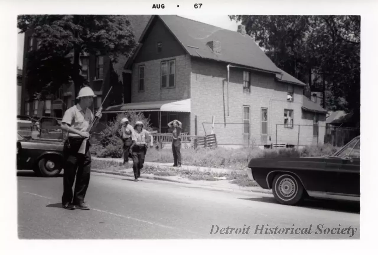 "One digital scan of a black and white photograph which shows a young white man being stopped by police during the 1967 Detroit Riot. The man is standing on a sidewalk on a residential street and has his hands on his head. Two police officers are walking away from him and another office is standing in the street and is holding a rifle. A police car is parked on the street in the left foreground and a car is parked in the right foreground. A house and vacant lot can be seen in the background."