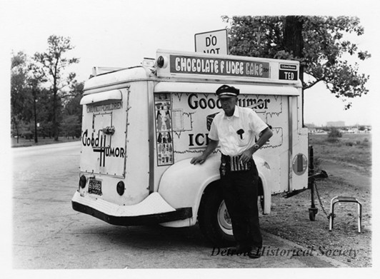 "Black and white photographic print mounted on board depicting a Good Humor Ice Cream trailer on Belle Isle, with a worker wearing a money changer standing in front. Labels on the trailer read, 'Chocolate Fudge Cake 30¢', 'Caution Children', and 'Fruit Stix, Raspberry 15¢'."