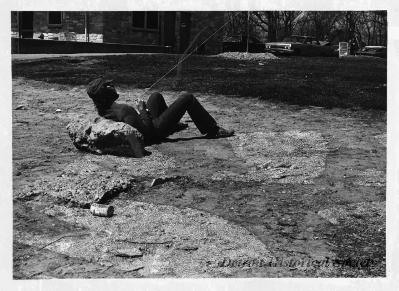 "Black and white photographic print depicting a man sitting on the ground with his back against a rock, flying a kite on Belle Isle. In the background are parked cars. In verso "WABX Kite-In". Dated April 15, 1973."