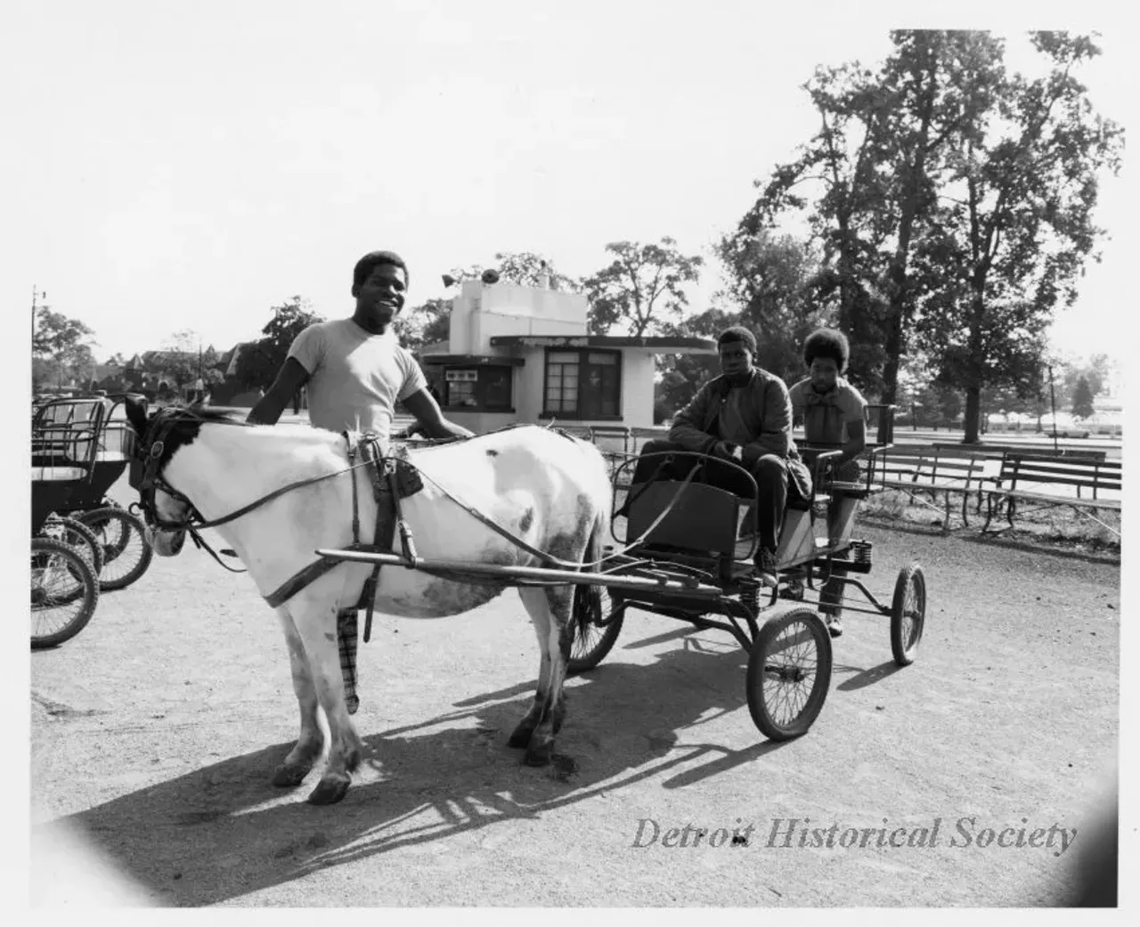 "Black and white photographic print depicting three young African American men in and around a pony cart on Belle Isle, with the stable and Conservatory in the background. Dated October 1973."