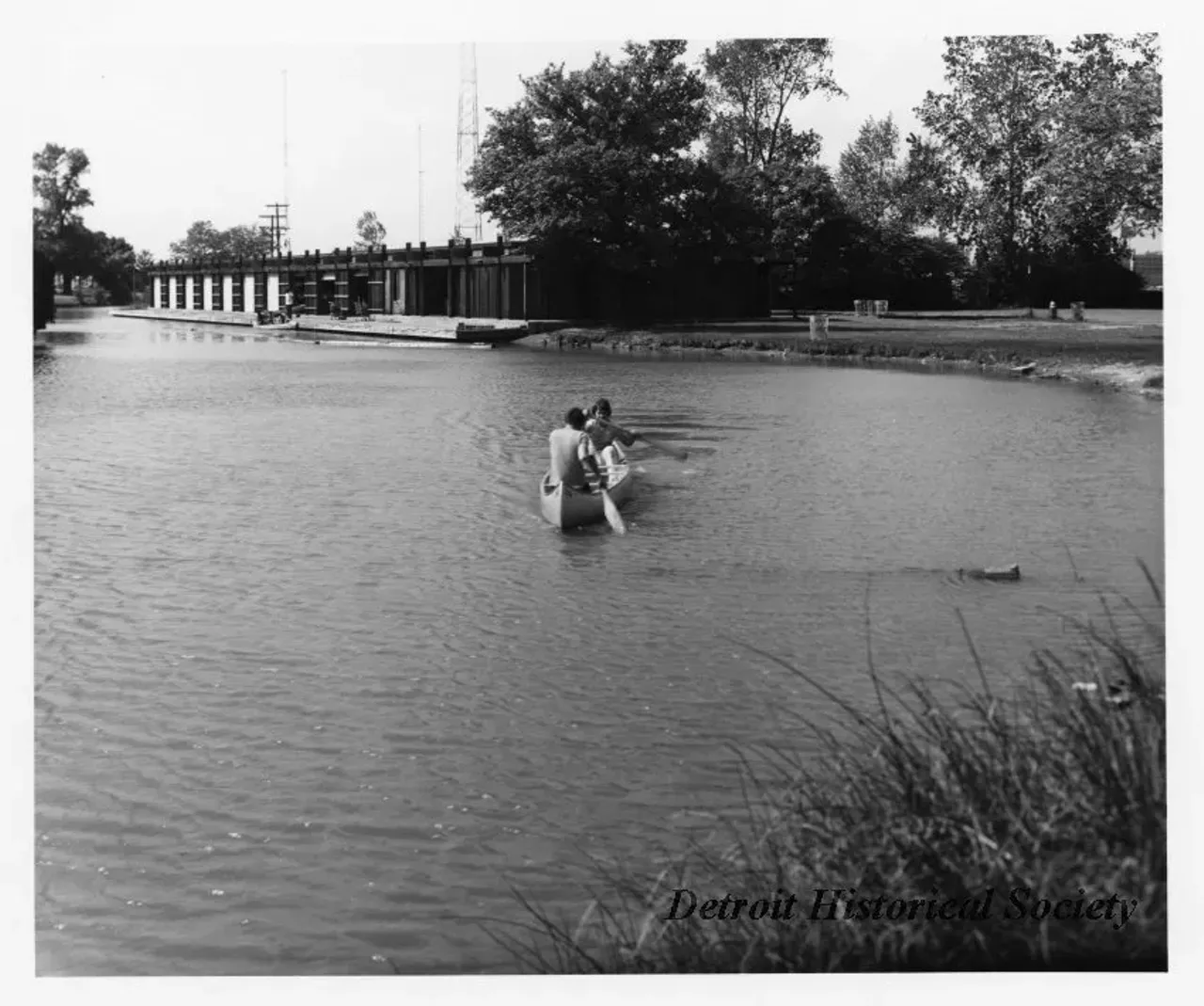 "Black and white photographic print depicting two people paddling a canoe through a canal on Belle Isle, with the Canoe Livery, trash cans, and radio towers in the background. Dated October 1973."