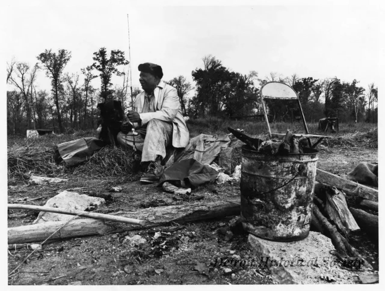 Black and white photographic print depicting African American man on Belle Isle holding fishing gear and accompanied by a dog. A metal folding chair and duffel bag are visible in background. Wood in a metal bucket is visible in the foreground. Dated October 1973.
