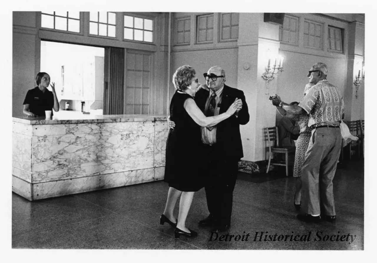 "Black and white photographic print depicting senior citizens dancing inside the Belle Isle Casino. A woman stands behind a marble counter, holding a cigarette. Dated November 1973."