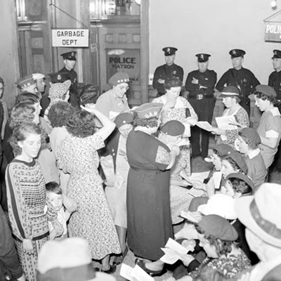 June 7, 1937: In a one-day general strike called by U.A.W. in Lansing, a group of people gather in an unidentified city building, with signs for Police and Garbage departments in the background, and uniformed police officers standing against the back wall, one man and one woman wear hats with "UAW" on them and many people appear to be reading aloud from pamphlets.