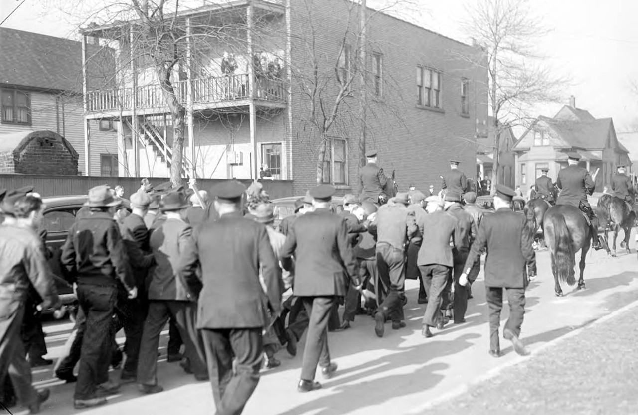 
March 30, 1938: A group of men walk in the street, escorted to the Federal Screw Works plant in Detroit by uniformed police officers on foot and mounted, with houses in the background.