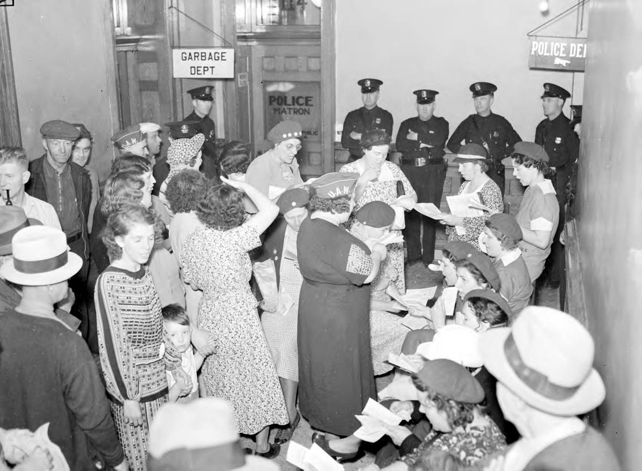 
June 7, 1937: In a one-day general strike called by U.A.W. in Lansing, a group of people gather in an unidentified city building, with signs for Police and Garbage departments in the background, and uniformed police officers standing against the back wall, one man and one woman wear hats with "UAW" on them and many people appear to be reading aloud from pamphlets.