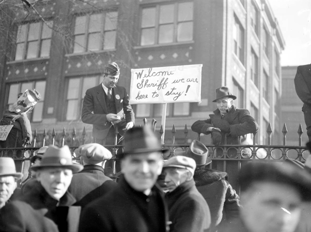 
March 17, 1937: Men gather outside the Dodge plant in Detroit during a sit-down strike. One man stands next to a picket sign that reads, "Welcome Sheriff, we are here to stay!"
