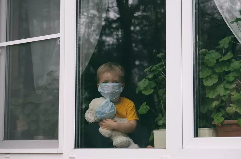 Sad child in protective medical masks looks out the window. - Shutterstock