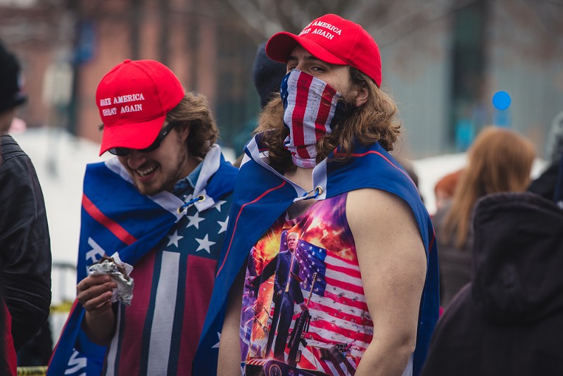 Two Trump supporters sporting MAGA hats, draped in Trump flags. - Shutterstock
